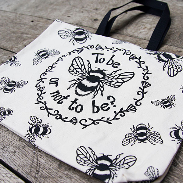 Tote bag made of heavyweight unbleached cotton canvas with mid-length black webbing handles. The bag is printed with a black design adapted from an original print of a bumble bee with a striped body and paneled wings, and a quote from Shakespeare play, Hamlet (To be or not to be?). In a ring around the bee and the quote are flowers and leaves. Outside this main design are a number of smaller bees flying in different directions.