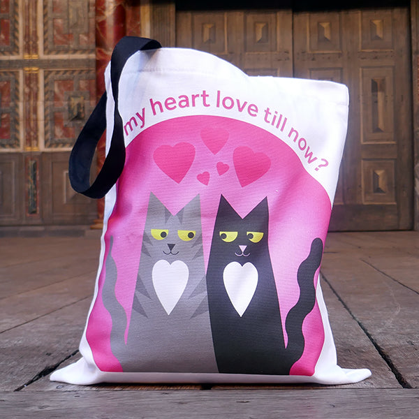 White cotton bag with black mid-length handles. A large print is on both sides of two cartoon cats, one a grey tabby , the other black. Both cats are smiling and looking lovingly at each other, both have a heart shaped white chest patch. They sit together in front of a pink circle with love hearts above them. Around the circle is a quote from Shakespeare play, Romeo & Juliet, "Did my heart love till now?" in pink letters.