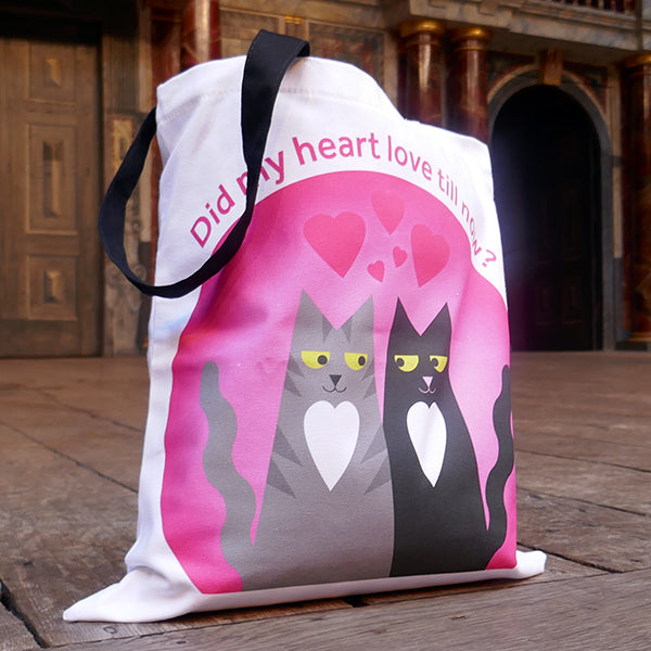 White cotton bag with black mid-length handles. A large print is on both sides of two cartoon cats, one a grey tabby , the other black. Both cats are smiling and looking lovingly at each other, both have a heart shaped white chest patch. They sit together in front of a pink circle with love hearts above them. Around the circle is a quote from Shakespeare play, Romeo & Juliet,