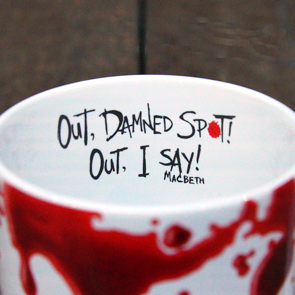 White earthenware coffee mug with a design of realistic red blood drips circling the rim and dripping down the mug. Inside the inner rim of the mug is a quote from Shakespeare play, Macbeth (Out, damned spot! Out, I say!) written in angry hand-drawn black letters. The O in the word spot is represented by a single drop of red blood.