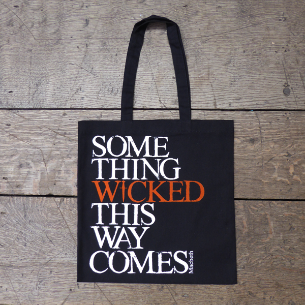 Black cotton bag with mid-length black handles printed with a quote from Shakespeare play, Macbeth (Something wicked this way comes). The lettering is bold serifed capital letters which are slightly distressed printed in white, except for 'WICKED) which is printed in orange. The 'I' of wicked is replaced by a dagger.