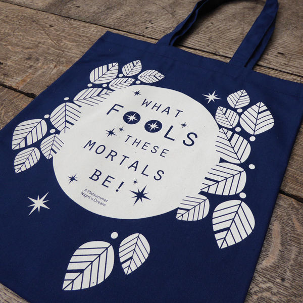 A  cotton book bag for Shakespeare's Globe. The bag is midnight blue with mid-length matching handles. Printed on the bag in white is a design consisting of a white circle (to represent the full moon) surrounded by stylised white leaves and stars. Across the face of the moon is a quote from Shakespeare play, A Midsummer Night's Dream (What fools these mortals be!) in midnight blue. The lettering is slim capitals.