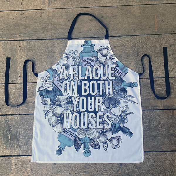 White cotton apron with black neck loop and ties, printed with an image celebrating Shakespeare play, Romeo & Juliet. A background of black and white flowers and green poison bottles with a quote fro the play, "a plague on both you house" printed in white capital letters.