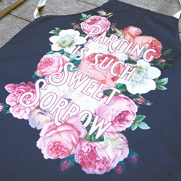 Black cotton apron with natural ties and neck loop printed with an image celebrating Shakespeare play, Romeo and Juliet. A photographic background of cream and peach roses, over this a quote from the play, "parting is such sweet sorrow" in fancy light pink capital letters with a darker pink outline