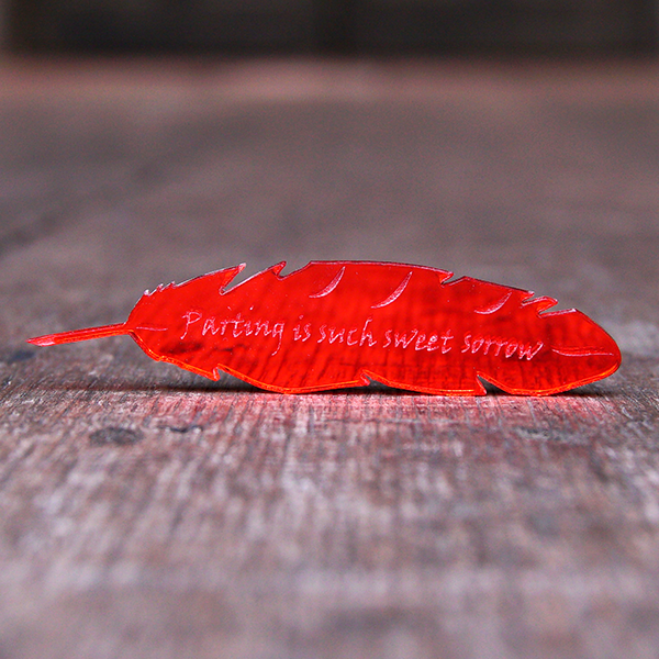 Red mirrored acrylic brooch shaped like a quill and engraved with a quote from Romeo and Juliet