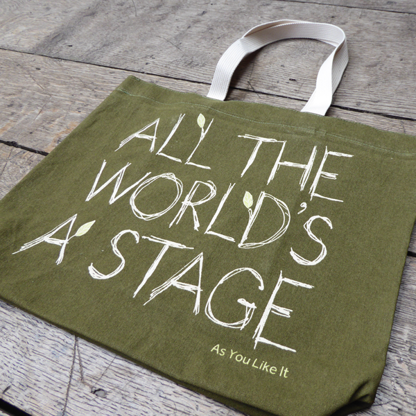 Sturdy sage green tote bag with a quote from Shakespeare play As You Like It (all the world's a stage) printed in white. The lettering is in a hand-drawn scribbled style to represent woodgrain and several of the letters have little lime green leaves growing out from them. The title of the play is printed in lime green under the quote. The bag has white handles.