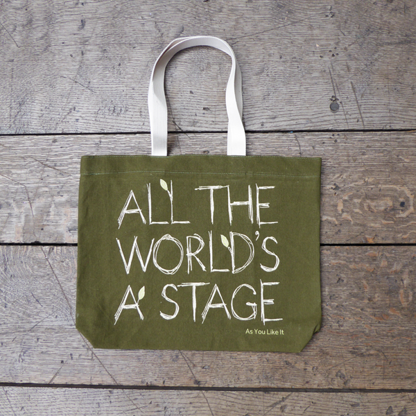 Sturdy sage green tote bag with a quote from Shakespeare play As You Like It (all the world's a stage) printed in white. The lettering is in a hand-drawn scribbled style to represent woodgrain and several of the letters have little lime green leaves growing out from them. The title of the play is printed in lime green under the quote. The bag has white handles.