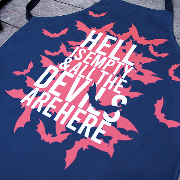 Black cotton apron with black ties and neck loop, printed with a design celebrating Shakespeare play, The Tempest. A cloud of red bats emanate from the centre of the apron. Over the top of the bats is a quote from the play (hell is empty and all the devils are here) in white capital letters.