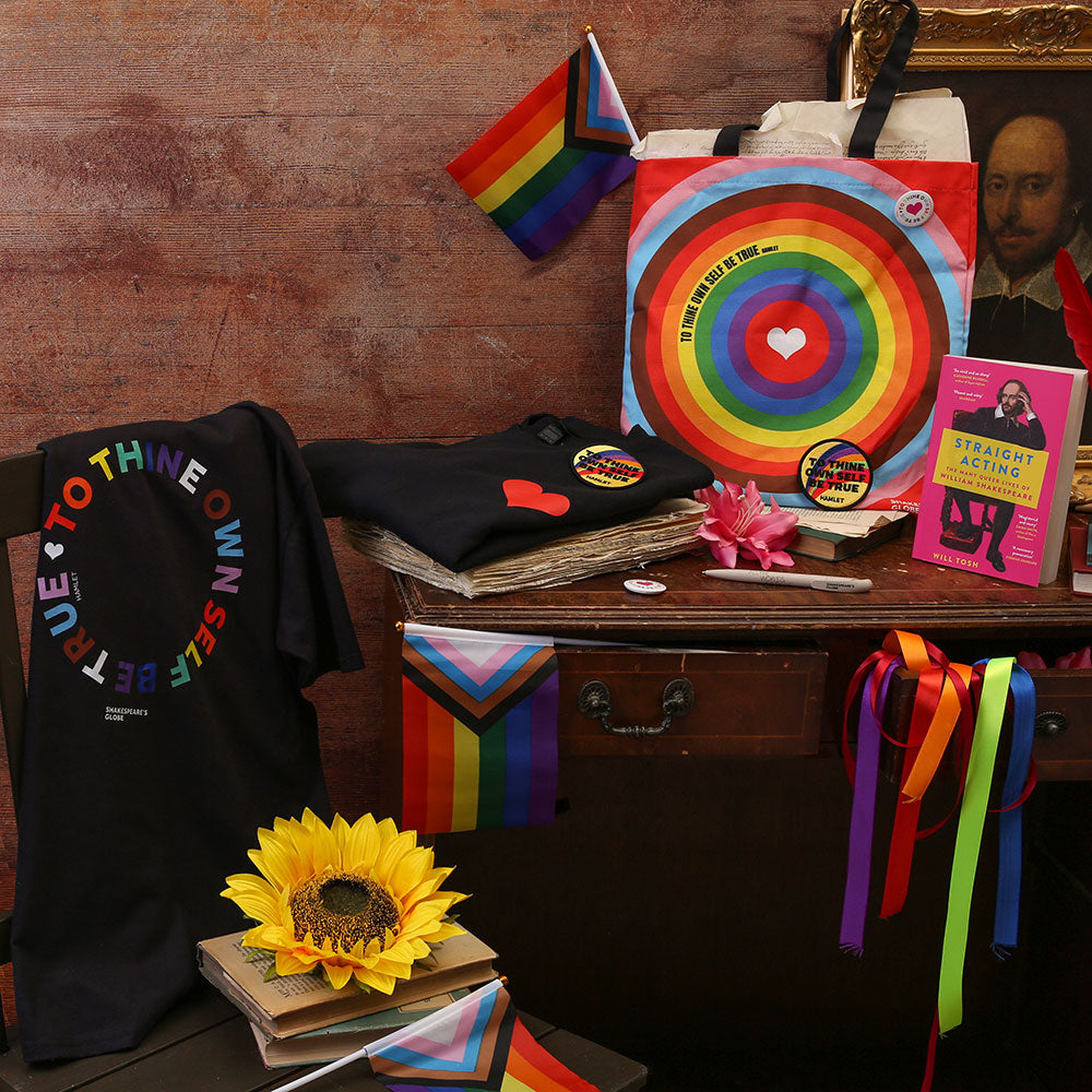 A white t-shirt, a tote bag, a sew on patch and a boopk with 'Love' printed on it in red, pink, blue and green sits on a dark wood background with colourful ribbon
