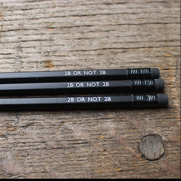 Three black pencils with '2B OR NOT 2B' text on a wooden surface