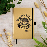 Notebook with 'Though she be but little, she is fierce' quote on a wooden surface with plants.