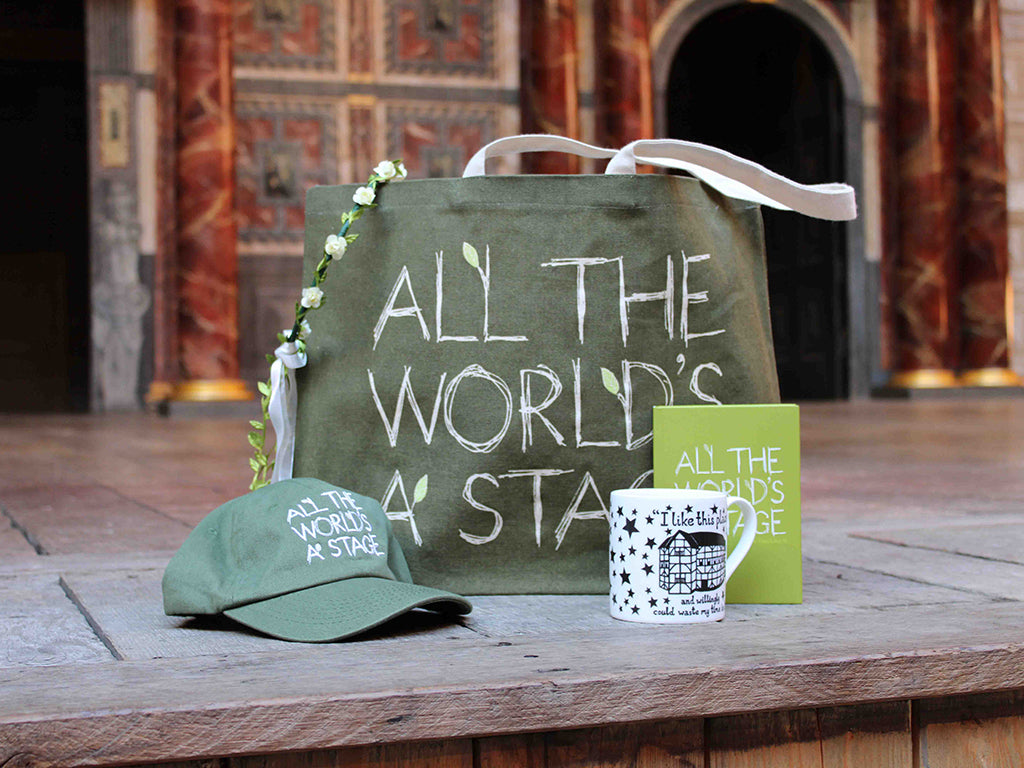 Wood panel stage floor with As You Like It sage green colour merchandise, left from right, baseball cap, paperback manga, tote bag, leaf brooch, hardback journal, purse