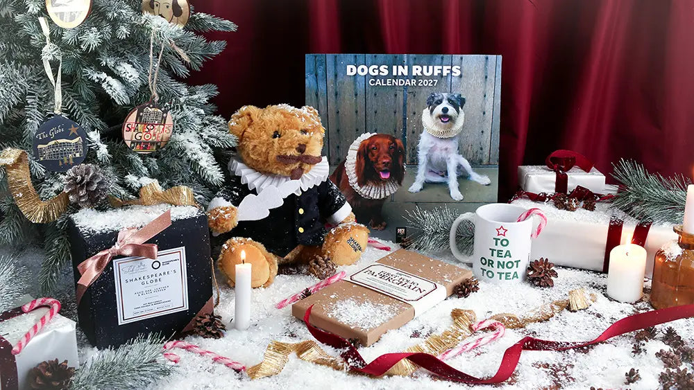 Festive Shakespeare-themed Christmas display featuring a Shakespeare teddy bear in period costume, Dogs in Ruffs 2027 calendar, ‘To Tea or Not to Tea’ mug, Shakespeare’s Globe candle, and First Folio paperchain kit, surrounded by a decorated tree, wrapped gifts, candles, candy canes, pinecones, ribbons, and artificial snow.