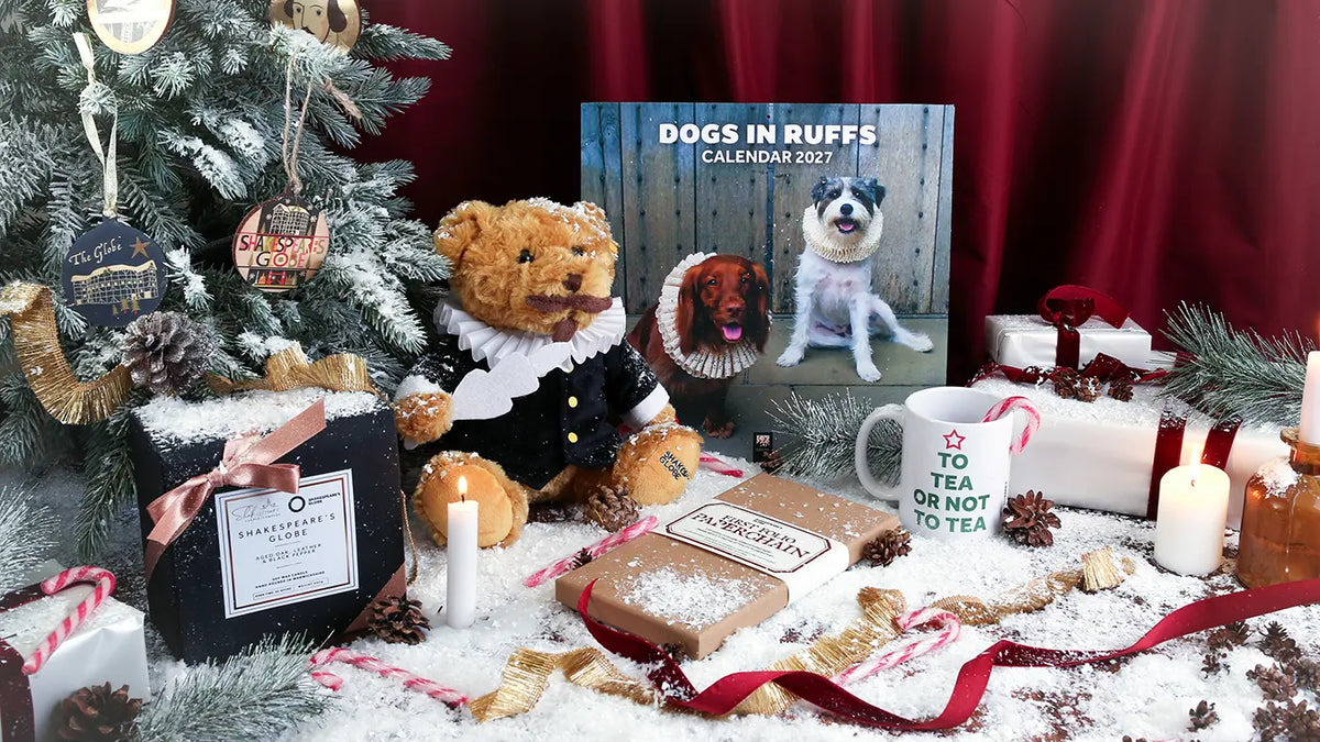 Festive Shakespeare-themed Christmas display featuring a Shakespeare teddy bear in period costume, Dogs in Ruffs 2027 calendar, ‘To Tea or Not to Tea’ mug, Shakespeare’s Globe candle, and First Folio paperchain kit, surrounded by a decorated tree, wrapped gifts, candles, candy canes, pinecones, ribbons, and artificial snow.