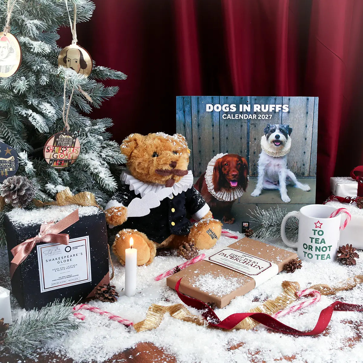 Festive Shakespeare-themed Christmas display featuring a Shakespeare teddy bear in period costume, Dogs in Ruffs 2027 calendar, ‘To Tea or Not to Tea’ mug, Shakespeare’s Globe candle, and First Folio paperchain kit, surrounded by a decorated tree, wrapped gifts, candles, candy canes, pinecones, ribbons, and artificial snow.