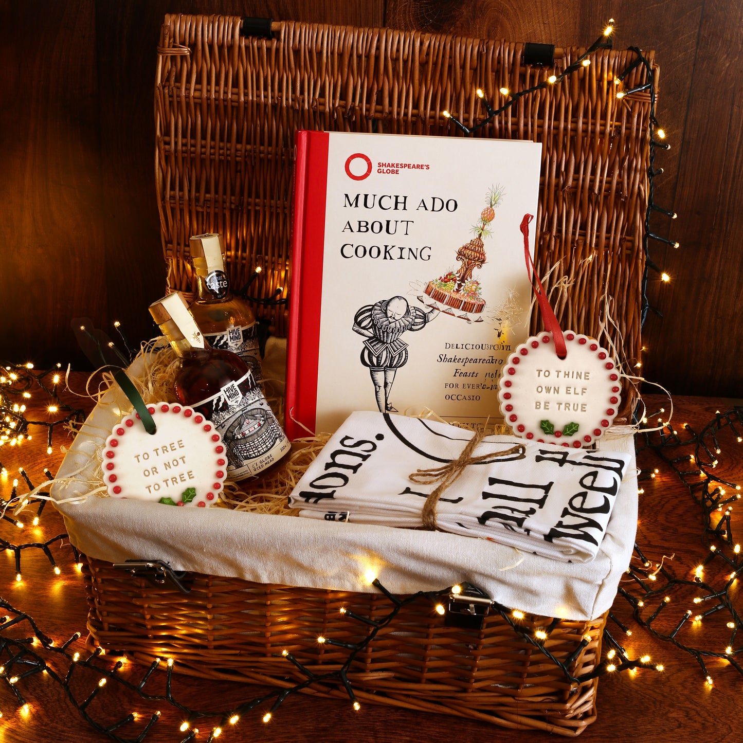 wicker hamper full of biscuits, mead, a cookbook and a tea towel on a wooden surface with fairy lights