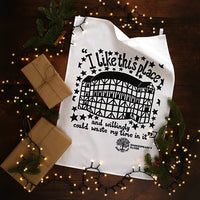 White tea towel with black text and design on a wooden surface with Christmas decorations.