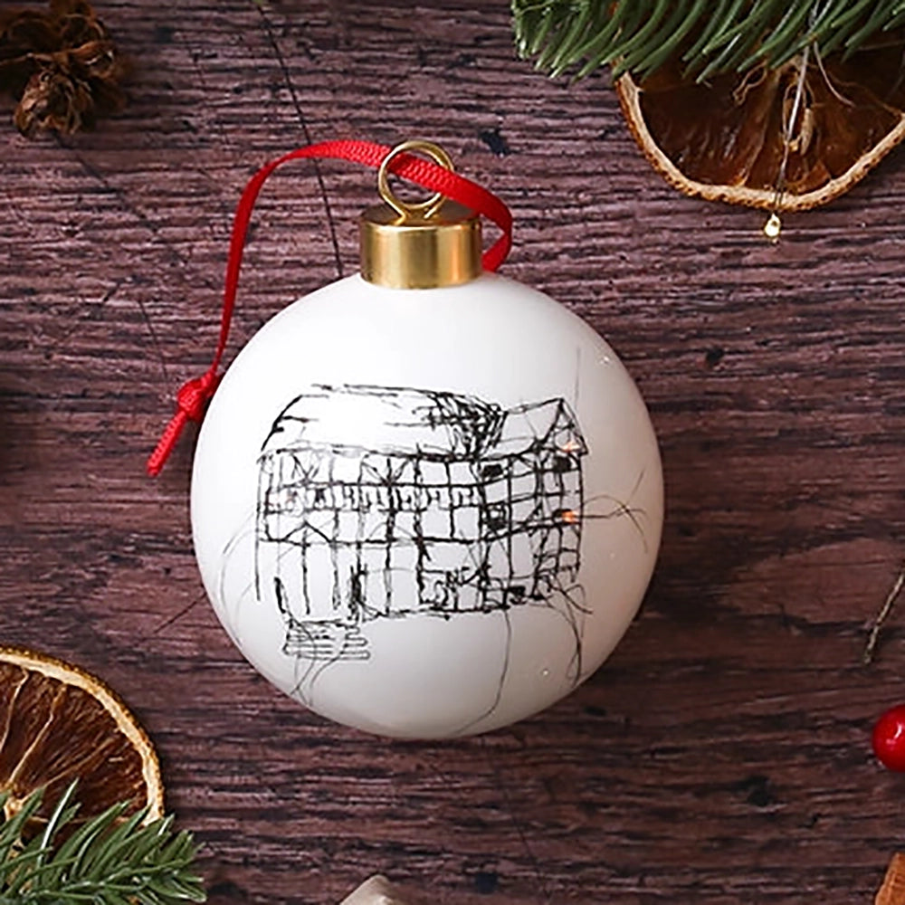 White glass bauble with black etched drawing of Shakespeare's Globe and gold top, with red tied ribbon, on wooden background with Christmas greenery, candles, orange slices and wrapped brown paper packages 