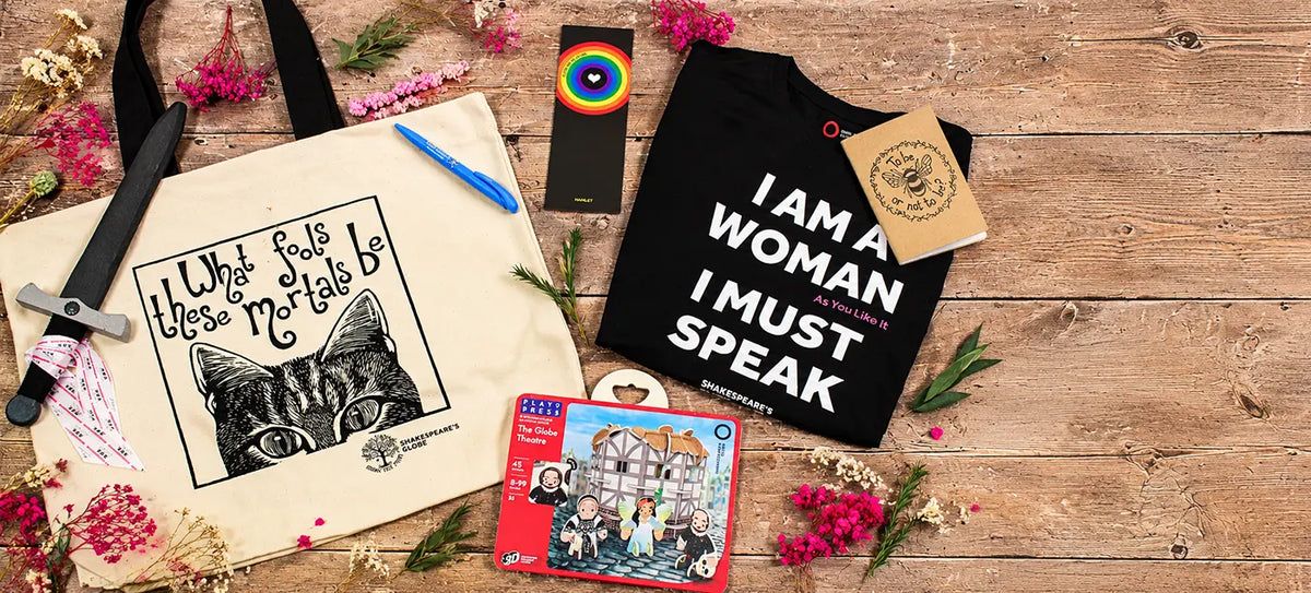 A styled flat-lay of multiple items on a rustic wooden surface: a cream canvas tote bag with a black cat illustration and the text 'I What Feels Most True To Me Is', a black T-shirt with large white text reading 'I AM A WOMAN I MUST SPEAK' and a tan hangtag, a small rectangular rainbow item, a comic book or card with a colorful illustrated cover, and a black pen, all arranged with dried flowers and greenery.