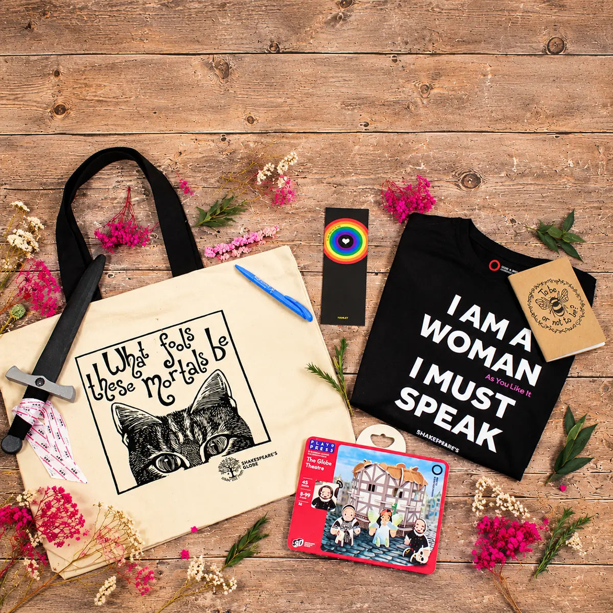 A styled flat-lay of multiple merchandise items on a rustic wooden background, including a beige tote bag with a black cat graphic and text, a black T-shirt with bold white text, a small colorful card or book, a notebook, a sword prop, and scattered pink flowers.