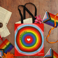 Colourful red bag with black handles and a design featuring all the colours of the Progress Pride flag on a wooden surface