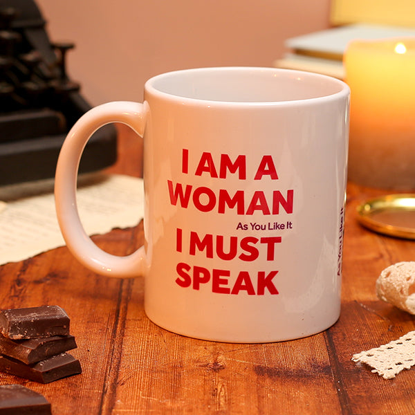 White ceramic mug with red text reading “I am a woman, I must speak,” displayed on a wooden desk with books and a candle