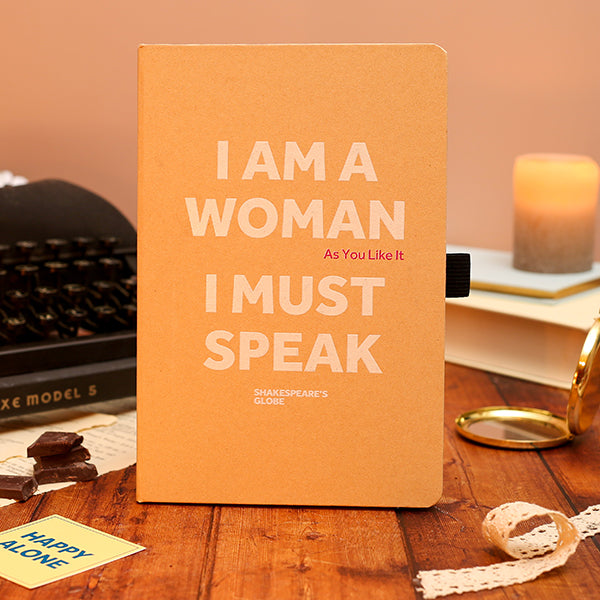 Notebook with 'I AM A WOMAN As You Like It I MUST SPEAK' text on a wooden surface with books and a candle.