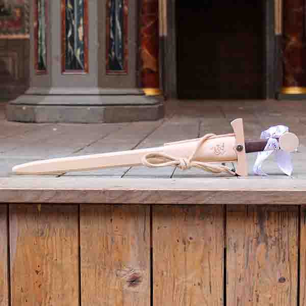 
                      
                        Wooden toy sword with a brown handle and a beige scabbard sitting on a wooden stage
                      
                    