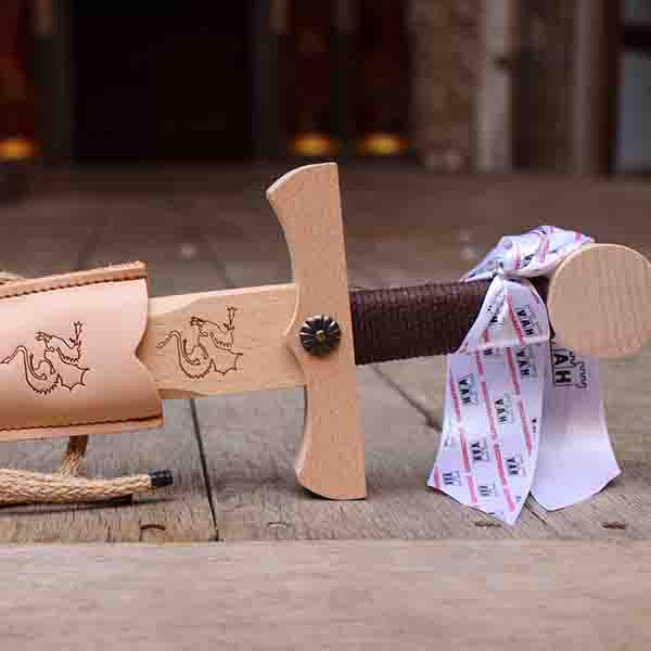 
                      
                        Wooden toy sword with a brown handle and a beige scabbard sitting on a wooden stage
                      
                    