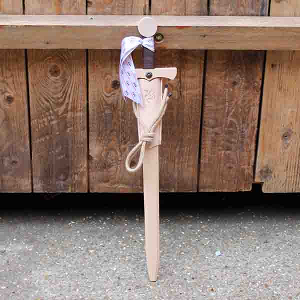 
                  
                    Wooden toy sword with a brown handle and a beige scabbard leaning against a wooden stage
                  
                