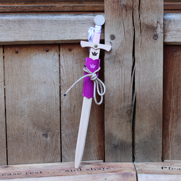 
                      
                        Wooden sword with a crown motif on the blade and a pink cord wrapped handle, with a pink felt half scabbard, leaning against a wooden post
                      
                    