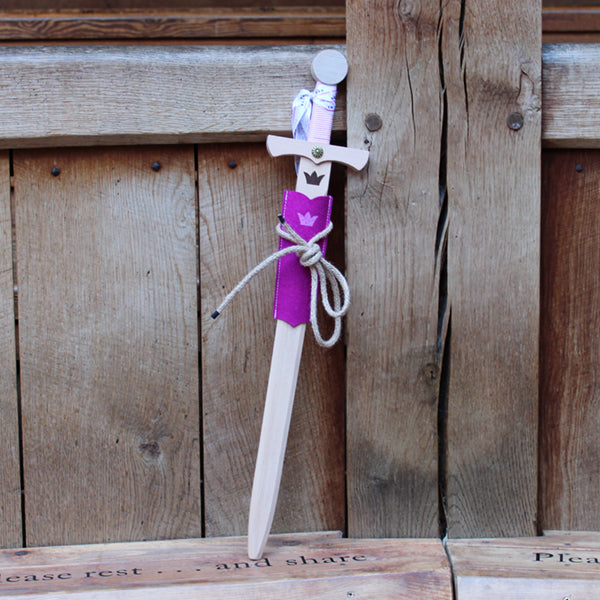 Wooden sword with a crown motif on the blade and a pink cord wrapped handle, with a pink felt half scabbard, leaning against a wooden post