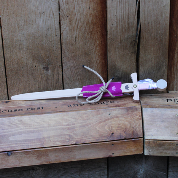 Wooden sword with a crown motif on the blade and a pink cord wrapped handle, with a pink felt half scabbard, leaning against a wooden post