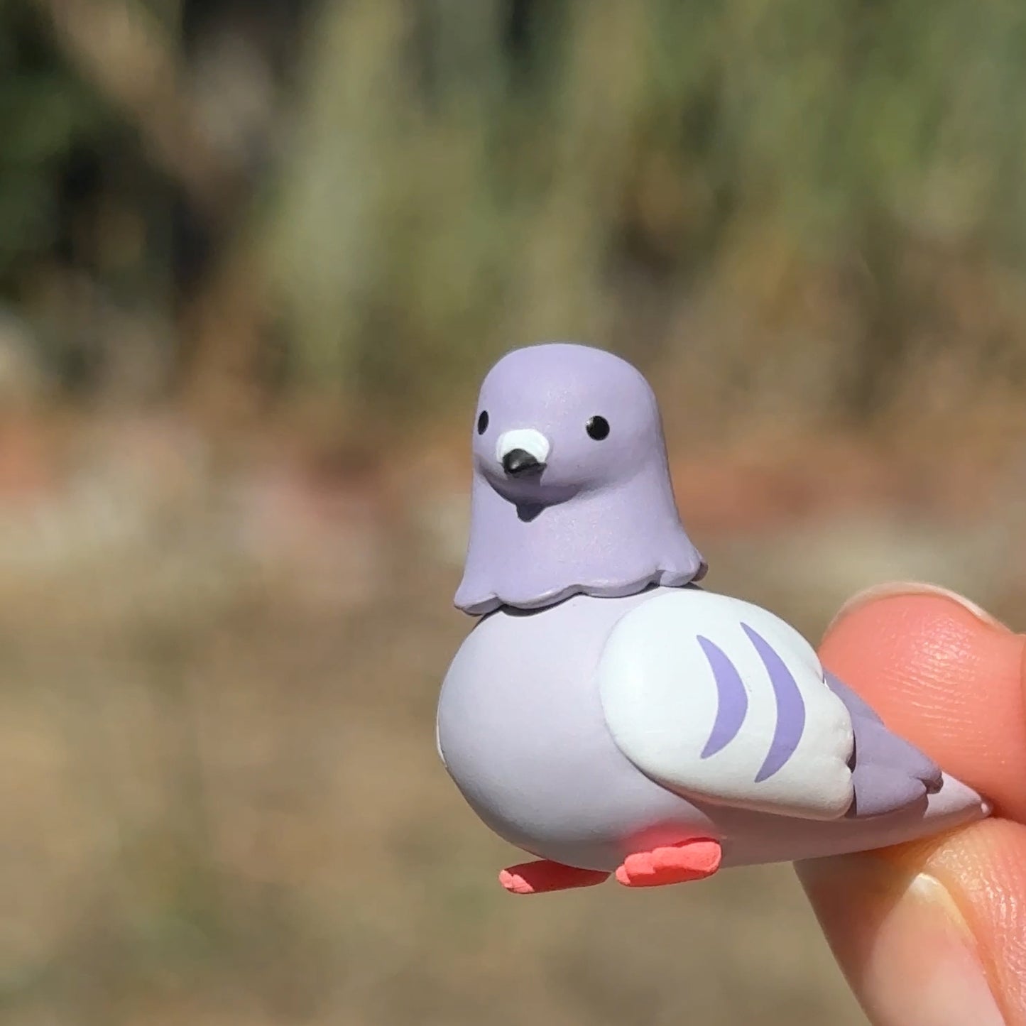 A small lavender and white pigeon figurine being held by a person's fingers against a blurred outdoor background.