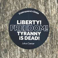 Round black sticker with white text on a wooden surface, featuring a quote from Julius Caesar.