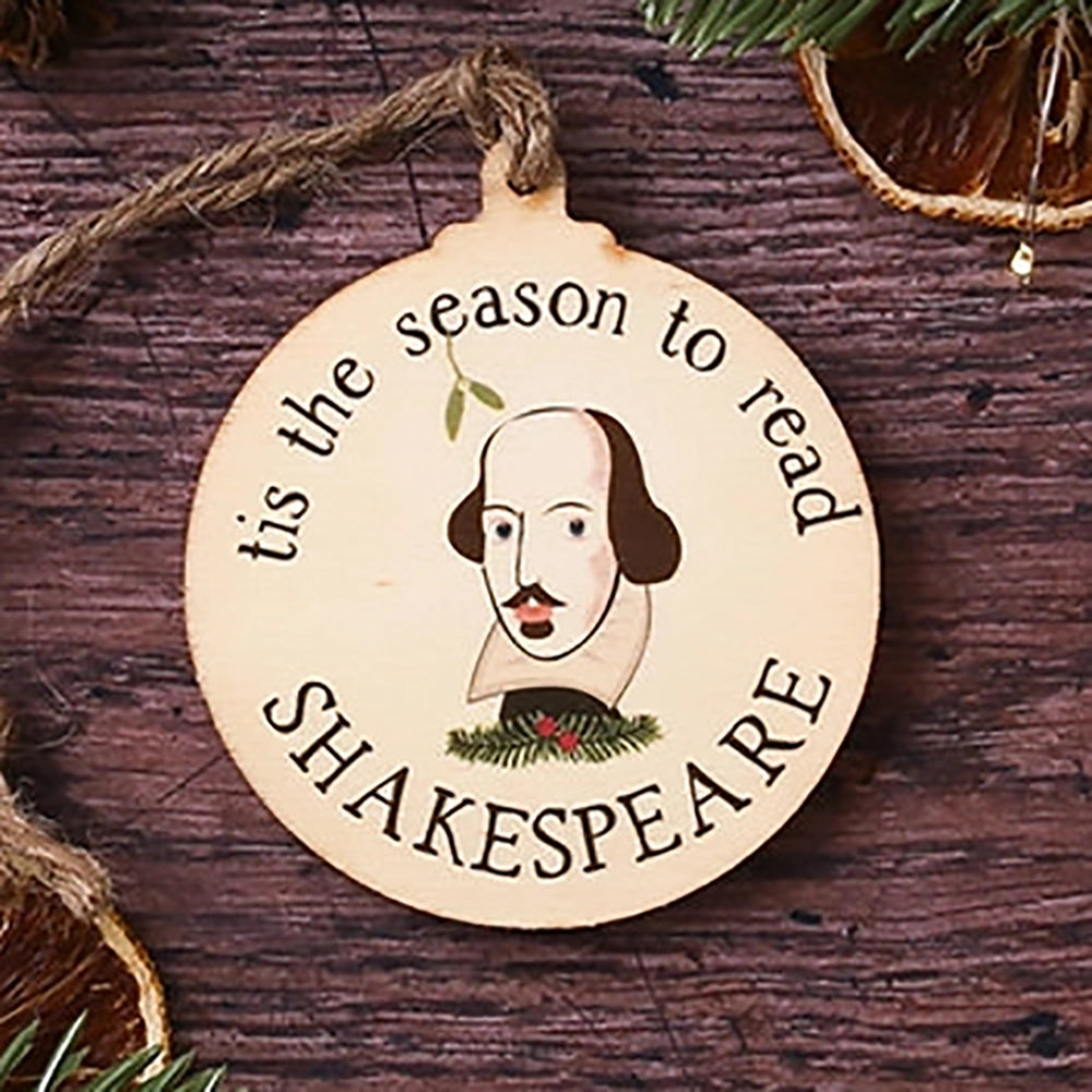 Round, flat, wooden Christmas decoration with Shakespeare's face in centre surrounded by 'tis the season to read' text., on wooden background with Christmas greenery