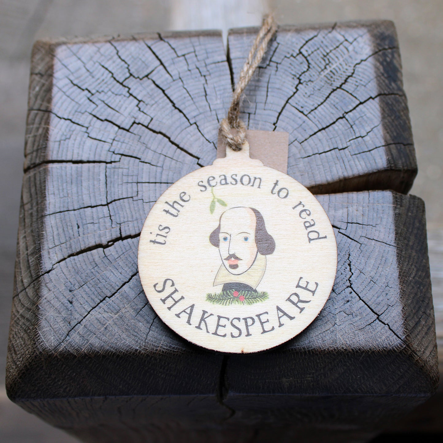 Round, flat, wooden Christmas decoration with Shakespeare's face in centre surrounded by 'tis the season to read' text.