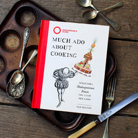 Cookbook titled 'Much Ado About Cooking' by Sam Bilton on a wooden surface with utensils.