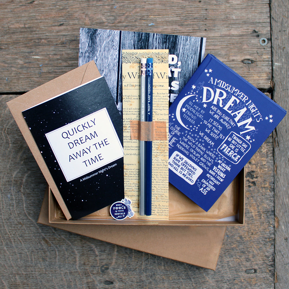 A kraft box, open with blue notebook, black greeting card, black mini print, blue and white pin badge and silver and blue pencil