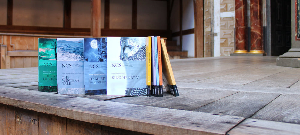 Paperback play texts standing up on wood panel stage, from left to right, teal, pale blue, blue, white, yellow, lilac, pale blue, orange, pale yellow