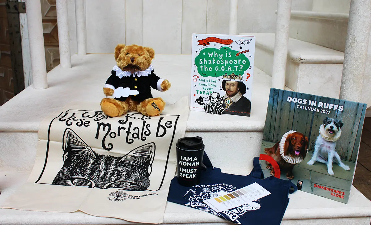 Shakespeare-themed gift items arranged on white stone steps, including a Shakespeare teddy bear, a cat-themed tea towel with a Shakespeare quote, a children's book about Shakespeare, a reusable coffee cup with the slogan "I AM A WOMAN I MUST SPEAK," a navy tote bag with a Shakespeare-inspired print, and a Dogs in Ruffs 2027 calendar featuring dogs dressed in Elizabethan ruffs.