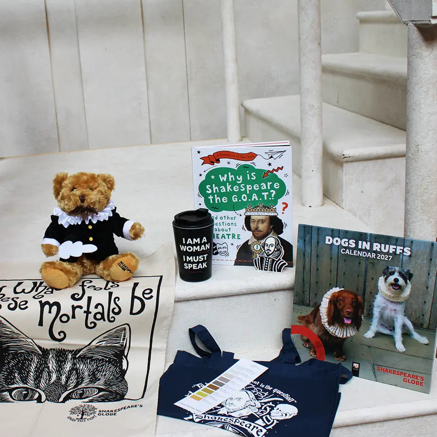 Shakespeare-themed gift items arranged on white stone steps, including a Shakespeare teddy bear, a cat-themed tea towel with a Shakespeare quote, a children's book about Shakespeare, a reusable coffee cup with the slogan "I AM A WOMAN I MUST SPEAK," a navy tote bag with a Shakespeare-inspired print, and a Dogs in Ruffs 2027 calendar featuring dogs dressed in Elizabethan ruff