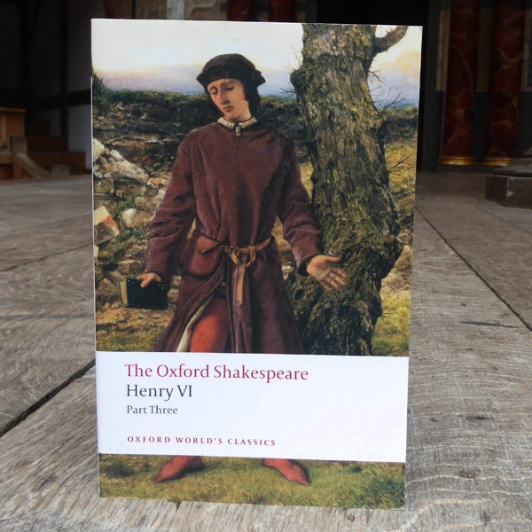 Paperback book on wood panel stage, book cover has man in brown robes, holding a black book, standing in front of a knobbly tree in a field