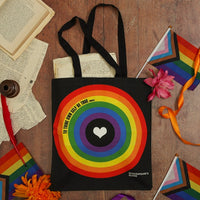 Black tote bag with rainbow circle design and heart, surrounded by books and rainbow-themed items on a wooden surface.