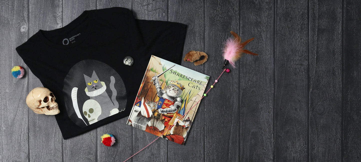 Grey wood panel background with small prop skull, folded black Shakespeare's cat t-shirt, paperback Shakespeare Cats book and scattered cat toys