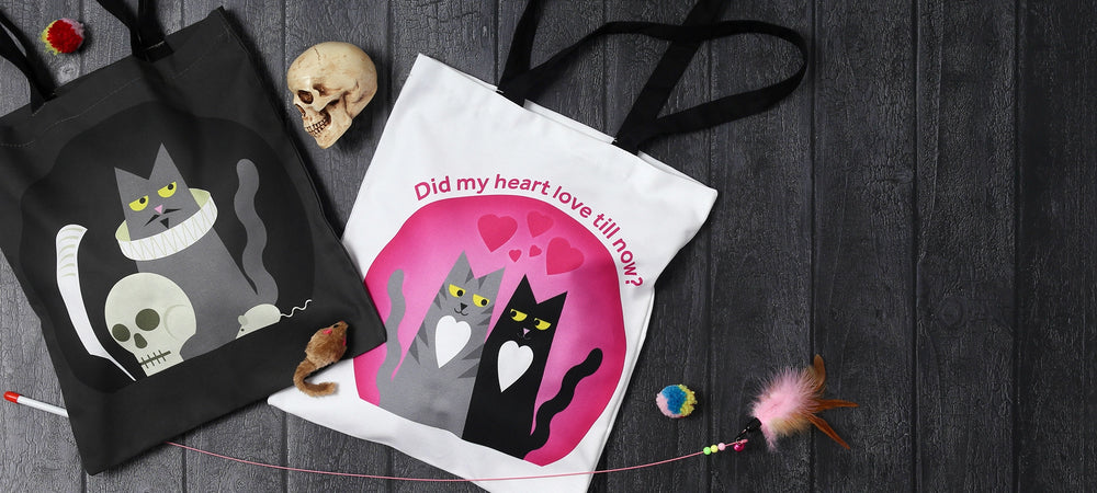 Two Shakespeare cats tote bags on grey wood panel floor, from left to right, grey Hamlet cat with skull, white and pink Romeo and Juliet cats bag with black handles