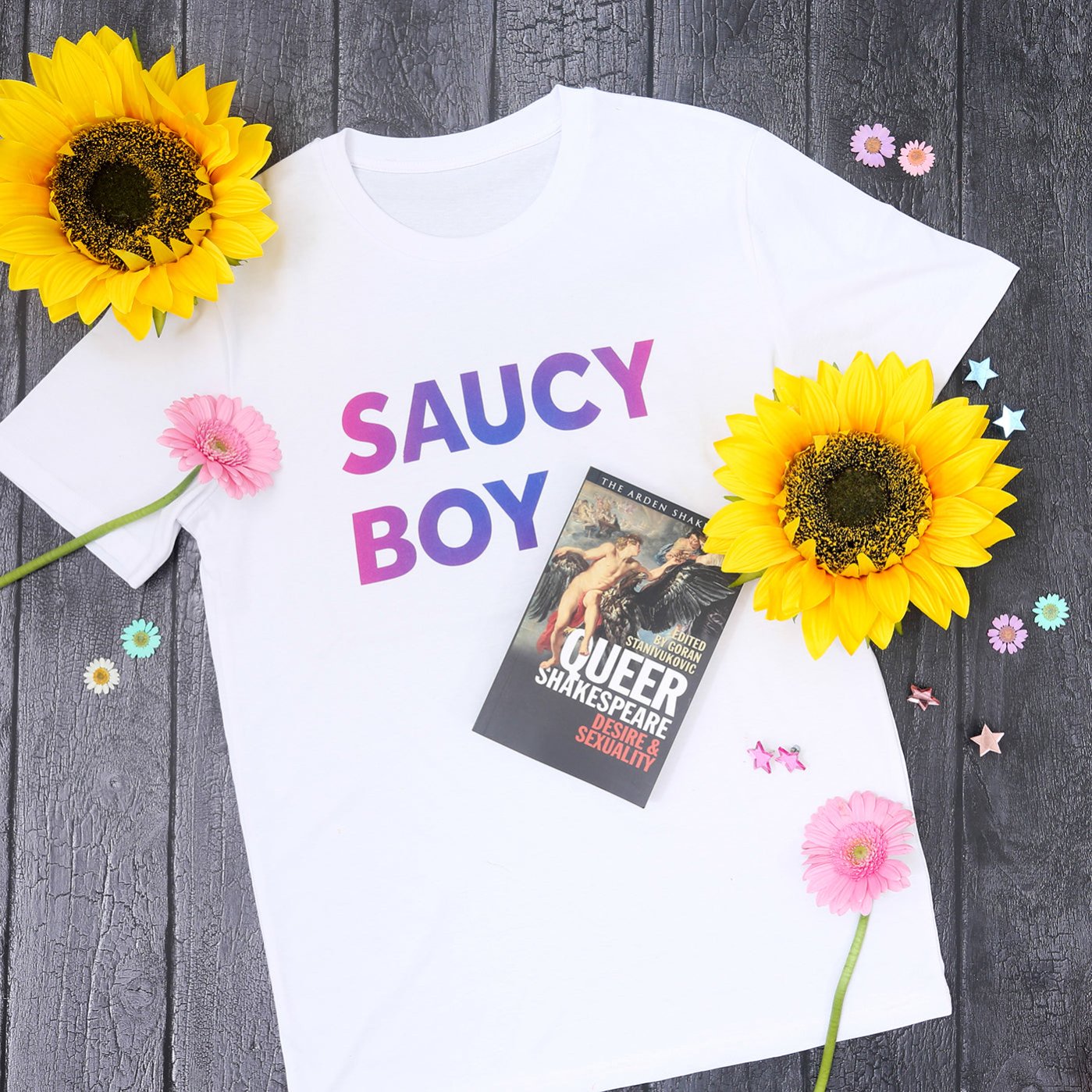 White cotton t-shirt with purple and pink gradient text on grey wood panel background, surrounded by sunflowers