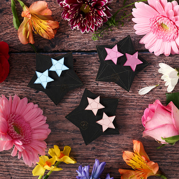 Pastel coloured star earrings on black backing card on wooden surface with flowers scattered about