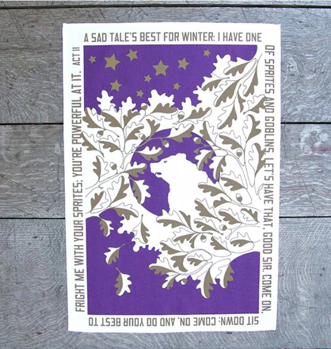 White cotton tea towel, portrait orientated, with purple and gold leaves surrounding polar bear design and gold text