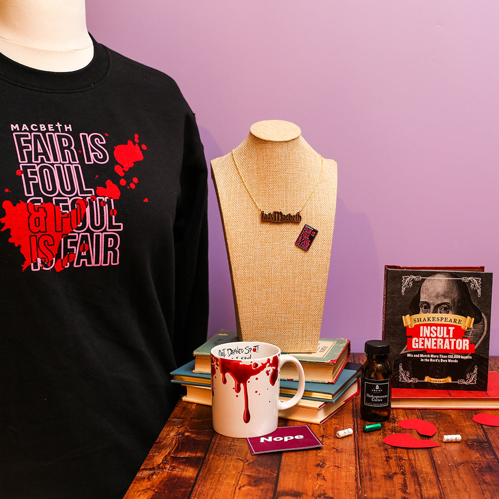Black sweatshirt with 'Macbeth' quote, mug with red liquid, books, and a necklace on a wooden table.